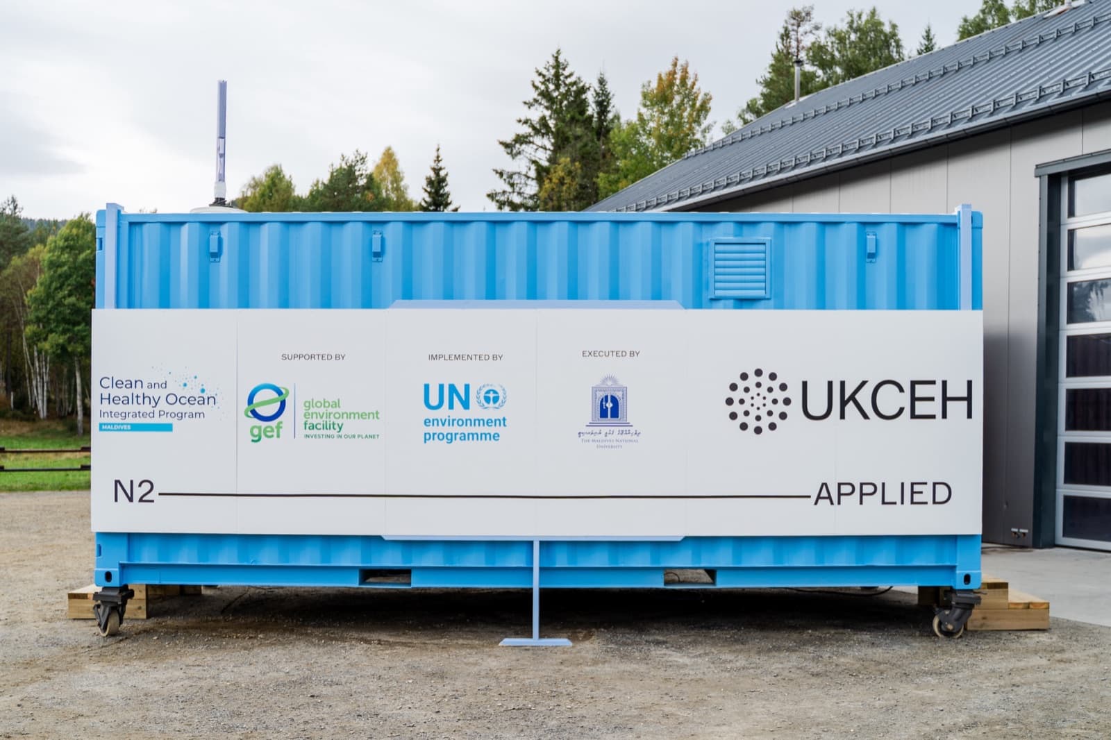 A pale blue containerised N2 unit on timber blocks outside a hall, its flank panelled with the Clean and Healthy Ocean programme's partner marks — GEF, UN Environment Programme and UKCEH — above the N2 Applied lockup